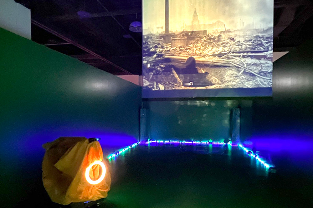 The contrasts between the robotic arm (covered with cloth on the left, it represents the presence of media in war), photos of Nagasaki in ruins projected on the screen, and ironic statement prompt reflection on mediated warfare.